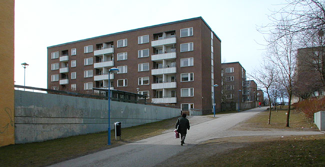 Gångväg norr om anläggningen. Betongmur vid terrassen framför stadsdelsnämndens hus. 
SAK05141 Sthlm Rinkeby, Kvarnberget 2, Rinkebyplan 2-8 (jmn nr) från ost












