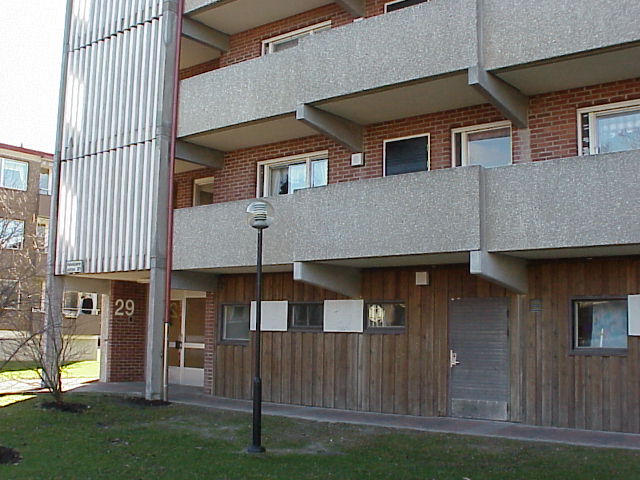 Närbild loftgång av betong med frilagd ballast, fasadbeklädnad i bottenvåning av träpanel och i övriga våningsplan av rött tegel. Entrén skyddas av en utbyggnad med vertikala ribbor.