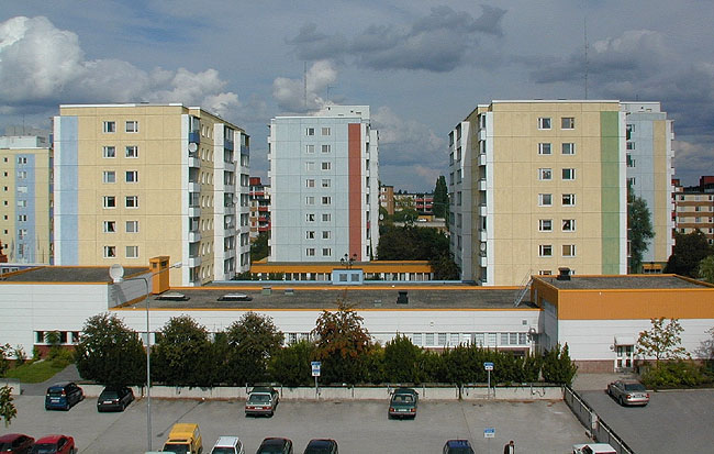 Höghusen är placerade på två parallella rader med en huskropps förskjutning. I förgrunden parkering och ett av de låga centrumbyggnaderna. 

SAK10354 Sthlm, Husby, Bergen 1, Ålesund 1, från SV
