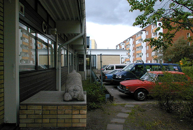 Parkering vid entrén mot Per Gyntgången. En smal grav närmast fasaden förser sammanbindningslängans källarvåning, dit biblioteket är förlagt, med dagsljus. 
SAK10070 Sthlm, Husby, Lofoten 4, Lofotengatan 32-34, från NO
