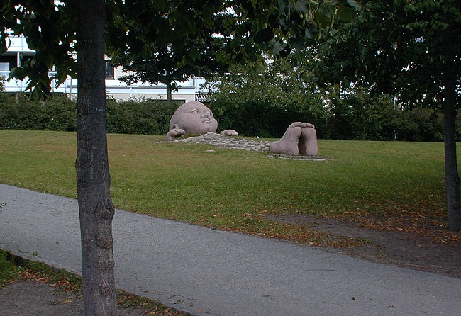 Skulpturen "Liten betraktare under blommigt täcke" av Nicke Rosén i sluttningen upp mot Thorvaldsengången. 
SAK12101 Sthlm, Kista, Själland 1, Själlandsgatan 150,152, från V