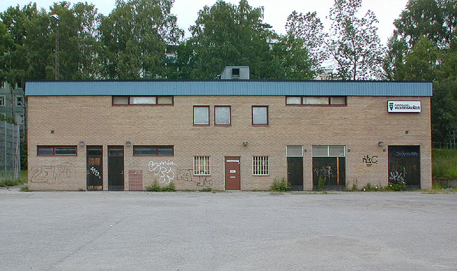 SAK10001 Sthlm, Husby, Akalla 4:1 1, nummer 85, Edvard Grieggången 1, från SO

Byggnaden är strängt kubisk. De flesta fönster och dörrar är asymmetrisk utplacerade på sydöstfasaden. 
