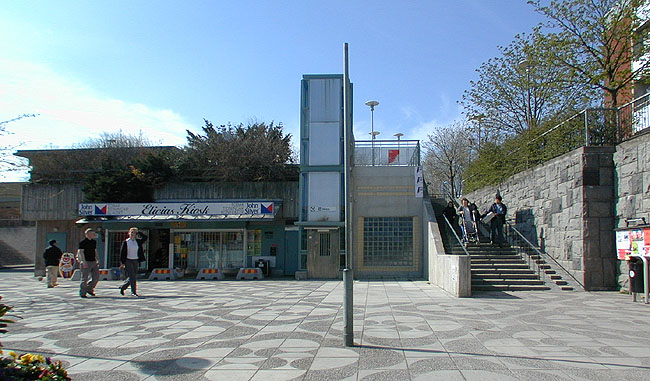 Tenstaplan med kiosk, hiss och trappa. Platsen ligger i samma nivå som bilvägarna. Till höger stödmur mot Tenstagången. 


SAK07416 Sthlm, Tensta Västra tunnelbaneuppgången, från sydöst




