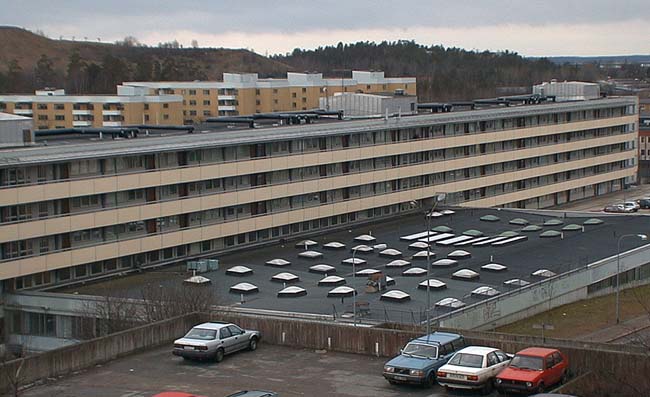 Sthlm, Vårberg, Vårholmen 4 Fjärdholmsgränd 4-38 (jmn nr) Vårbergsplan 3-11(udda nr) från nordost. Ett långsträckt loftgångshus är sammanbyggt med centrumhuset. 

