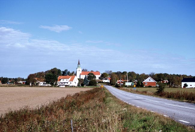 Kyrkan ligger på en mindre höjd omgiven av bebyggelse och med flacka åkrar runt om.
