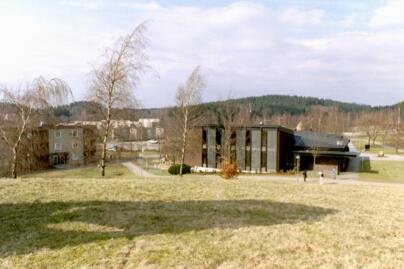 Hässleholmens kyrka och församlingshem ligger omgiven av olika bostadsområden och en skola till höger ur bild. Kyrkan ses här från kullen med klockstapeln, vars skugga faller över gräset.