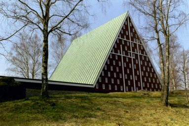 Sjöbo kyrka från 1962 ligger på en liten höjd med naturmark och ses här från parkeringen med sin karakteristiska korsprydda gavel översållad med små fönstergluggar.