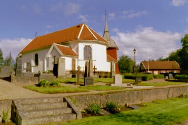 Brämhults kyrka med omgivande kyrkogård, klockstapel och det gamla skolhuset i bakgrunden, sett från sydöst. 