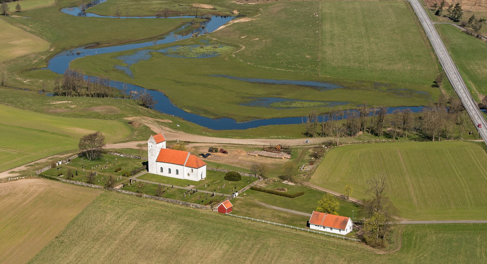 Flygbild över Lyngsjö kyrkoanläggning från sydöst med Vramsåns meanderslingor norr om kyrkan