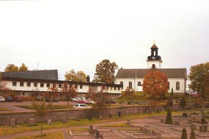 Församlingshemmet med kyrkan och del av kyrkogården. Foto från nordväst.
