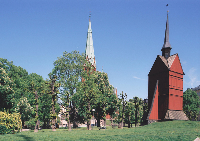 S:t Johannes kyrka, klockstapeln. Bakom grönskan skymtar kyrkobyggnaden med sitt torn.
